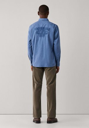 Hombre con camisa azul de manga larga con texto "FACONNABLE French Alps" en la parte trasera, pantalones marrones y zapatos marrón oscuro, de pie frente a una pared lisa.