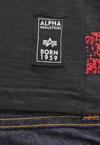 Svart tygmärke med "ALPHA INDUSTRIES" och "BORN 1959" i vitt, med ett rött grafiskt mönster och mörk denim med synlig gul söm.
