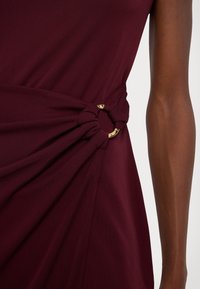 Burgundyklänning med draperat tyg och knutdetalj samt guldfärgad hårdvara vid midjan. Samt en mjuk textur och ärmlös design.