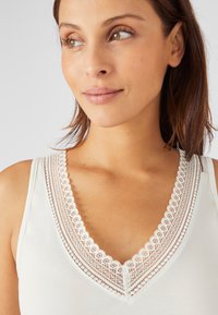 Camiseta blanca sin mangas con un borde de encaje alrededor del escote en V. Tela suave y un delicado detalle de borde ondulado que acentúa el escote.