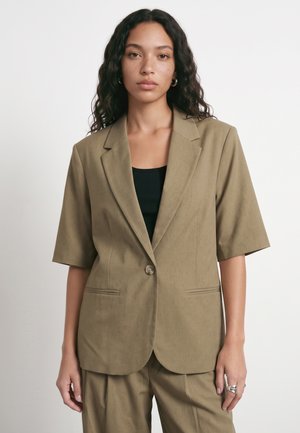 Jeune femme aux cheveux bouclés portant un blazer kaki à manches courtes et un pantalon assorti sur un haut noir, debout devant un fond uni.