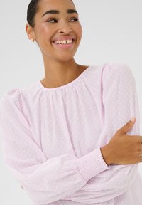 Blouse rose clair en tissu texturé avec de petits points en relief. Elle présente un col rond, des manches bouffantes et des poignets smockés.