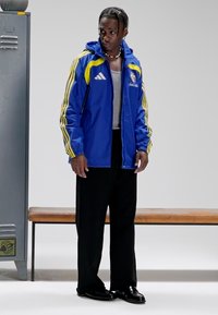 Blauwe waterdichte jas met gele strepen, grijs shirt, zwarte broek en glanzende zwarte schoenen. Staand naast een houten bank en kluis.