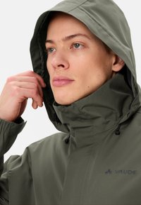 Groene waterdichte jas met capuchon, voorzien van een hoge kraag en elastische manchetten. Opvallend logo in donkerblauw op de borst.