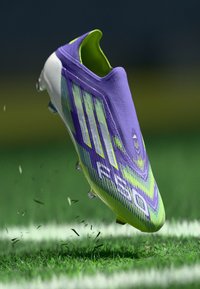 Lila Fußballschuh mit strukturiertem Obermaterial, das grüne Akzente und ein markantes Design aufweist. Schwebend über dem Gras, was Bewegung und Realismus zeigt.