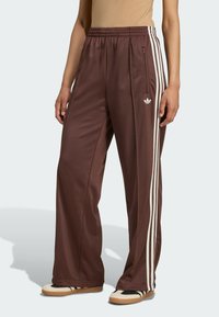 Pantalones deportivos de poliéster marrón con rayas blancas en los laterales, que cuentan con un bolsillo con cremallera y un logo bordado en la pierna izquierda. Diseño de pierna ancha.