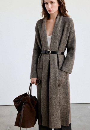 Femme portant un long manteau marron à chevrons ceinturé à la taille, tenant un grand sac en daim marron foncé, debout devant un fond uni.