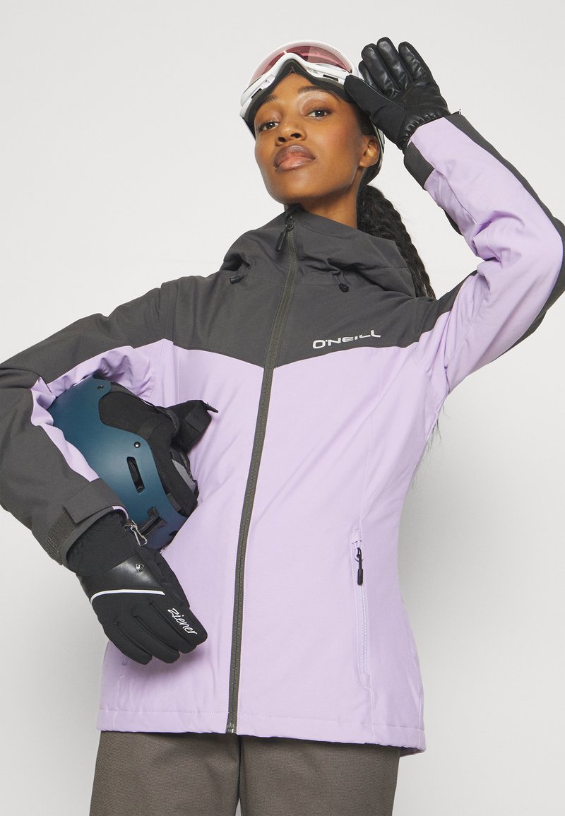 Lila und schwarze Skijacke mit Reißverschluss und Kapuze. Kombiniert mit schwarzen Handschuhen und einem blauen Helm, der sportliches Design und Funktionalität zeigt.