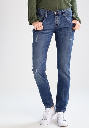 Osoba nosząca niebieskie, znoszone jeansy typu slim-fit z zapięciem na guziki, zielony długi rękaw oraz białe sneakersy, stojąca z rękami w kieszeniach.