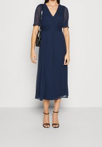 Marinblå midiklänning med v-ringning och korta genomskinliga ärmar, utrustad med ett figursytt midja och flödande kjol. Svarta remsandaler med klack och en liten svart väska.