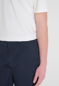 Pantaloni blu navy con una superficie liscia, caratterizzati da un sottile etichetta di marca. Presentati insieme a una maglietta bianca a maniche corte.