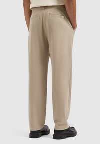 Beige, schmal geschnittene Hose mit seitlichen Taschen, hinteren Paspeltaschen und einer glatten Textur. Kombiniert mit schwarzem Schuhwerk.