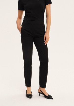 Kobieta ubrana w czarne dopasowane spodnie, czarną bluzkę z krótkim rękawem i czarne czółenka na obcasie z odkrytą piętą, stojąca na białym tle.