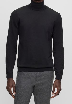 Mann trägt schwarzen Rollkragenpullover aus Langarmstoff und graue Wollhose, steht vor hellem, einfarbigem Hintergrund.