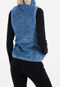 Blauer Kunstpelz-Weste mit hohem Kragen und weicher Textur, getragen über einem schwarzen Langarmshirt, mit einem kontrastierenden blauen Saum.