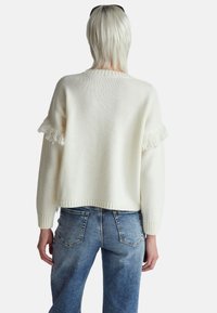 Biały sweter dziany z frędzlami na rękawach, ściągacz na dole i przy mankietach, w połączeniu z niebieskimi dżinsami. Widok z tyłu.