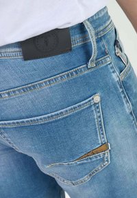 Jean en denim bleu avec une pièce en cuir à la taille, des poches arrière inclinées et des coutures dorées le long des coutures.