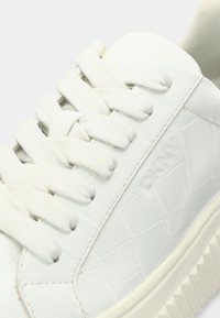 Λευκά συνθετικά αθλητικά παπούτσια με υφή κροκοδείλου, επίπεδα κορδόνια και λαστιχένια σόλα. Ανάγλυφο λογότυπο στο πάνω μέρος.