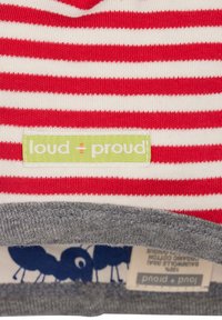 Tessuto a righe rosse e bianche con un'etichetta verde che legge "loud + proud". Orlo grigio con una stampa di formiche blu visibile sotto. Morbida trama di cotone.