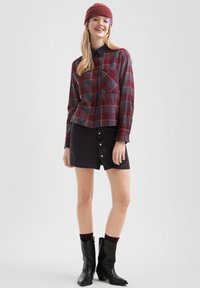 Jonge vrouw met een rode beanie, bril, geruit overhemd met knopen, zwarte minirok met knopen, zwarte sokken en zwarte middenlaarsjes, lachend.