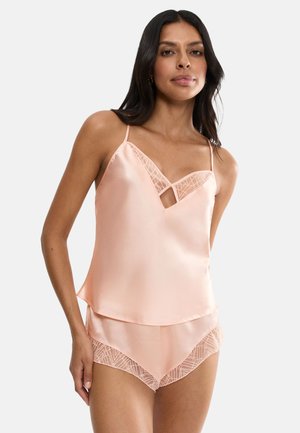Frau trägt hellrosa Satincamisole und passende Shorts mit Spitzenbesatz, steht vor einem schlichten hellen Hintergrund.