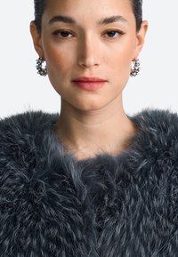 Donkergrijze faux fur jas met een pluizige textuur en een diepe halslijn. Het model draagt ronde oorbellen met parelachtige accenten.