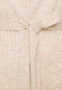 Närbild av ett beige stickat bälte knutet runt ett plisserat tygplagg.