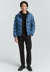 Blau gesteppte Daunenjacke mit Reißverschluss, Kinnschutz und zwei Fronttaschen. Schwarzes geripptes Sweatshirt und Hose mit schwarzen Schnürschuhen.