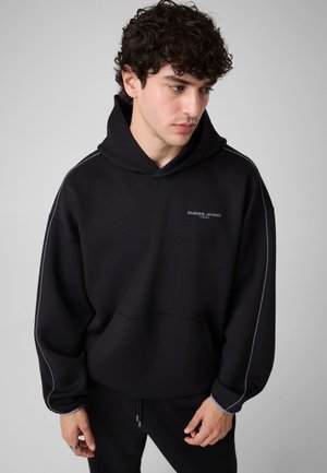 Junger Mann mit lockigem Haar trägt einen weiten schwarzen Hoodie mit dem Text "Guess Jeans 1981", schaut nach unten und steht vor einem schlichten, hellen Hintergrund.