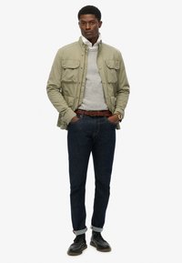 Olijfgroen lichtgewicht jack met meerdere voorzakken, grijze trui, donkere spijkerbroek, bruin gevlochten riem en zwarte leren schoenen.