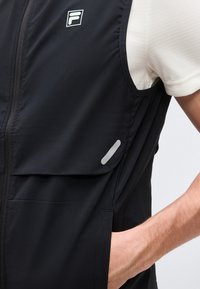 Άτομο που φοράει μαύρο γιλέκο χωρίς μανίκια με φερμουάρ, εμπρόσθια τσέπη με καπάκι, μικρή αντανακλαστική λωρίδα και λογότυπο στο στήθος πάνω από λευκό πουκάμισο με κοντά μανίκια.