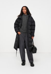 Casaco puffer preto com uma silhueta longa, apresentando fecho com zipper, punhos canelados e gola alta. Usado sobre um suéter cinza e calças escuras.