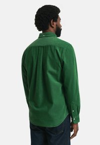 Chemise en velours côtelé vert avec col boutonné, détail de pli au dos et manches longues avec poignets à bouton. Tissu texturé et coupe ajustée.