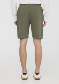 Pantaloni corti verde oliva con una vestibilità comoda, fascia elastica in vita e dettaglio in rilievo con il testo "EVERYONE" lungo il lato. Abbinati a scarpe sneakers bianche.