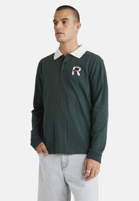 Green long-sleeve polo shirt with a white collar, featuring a floral logo patch on the chest and ribbed cuffs. Casual fit and soft fabric.
