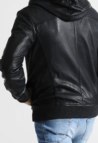 Giacca in pelle nera con cappuccio. Presenta polsini e orlo a coste, texture liscia e dettagli di cucitura discreti sulla schiena.