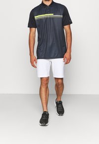 Marinblå pikétröja med ett gult rand- och prickmönster, kombinerad med vita shorts och svarta träningsskor. Avslappnad, sportig design.