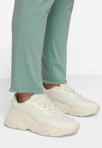 Zapatillas blancas con cordones, con una suela gruesa y parte superior texturizada, combinadas con pantalones verde claro que tienen dobladillos deshilachados y detalles de costura.