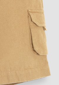 Beige pamut sort, textúrált felülettel és egy oldalsó zsebbel, melynek fedél van a tetején. Varrott szegély, tiszta befejezéssel.