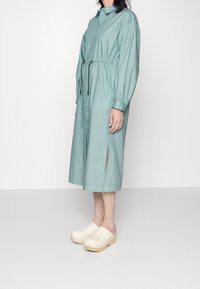 Donna che indossa un abito midi a righe blu chiaro con vita regolabile e spacchi laterali, abbinato a zoccoli in legno beige.