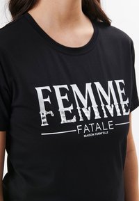 Sort bomulds T-shirt med et metallisk sølvgrafik, der står "FEMME FATALE" tværs over brystet. Kortærmet og med rund hals.