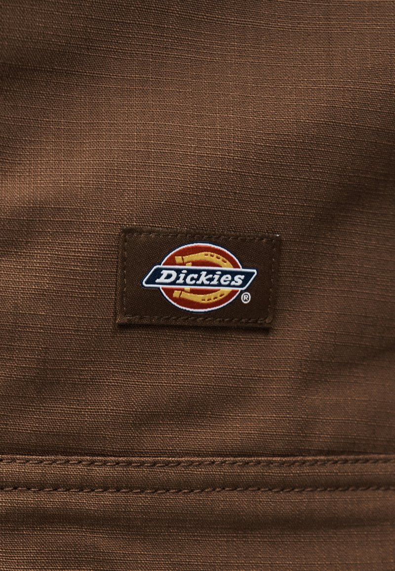 Tessuto marrone con un'etichetta rettangolare del marchio Dickies che presenta un logo a ferro di cavallo cucito sul materiale.