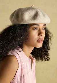 Világos bézs színű gyapjú barett, lekerekített formával és kis felső füllel. A modell ujjatlan rózsaszín kötött felsőt visel, bordázott textúrával.