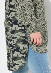 Grünes Langarmoberteil mit einem Leopardenmuster auf einer Seite und einem Camouflage-Muster auf der anderen. Verfügt über einen abgerundeten Saum und elastische Bündchen.