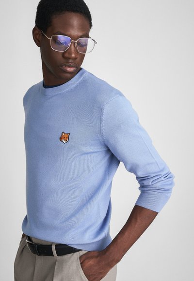 Pull en tricot bleu clair avec un col rond, présentant une tête de renard brodée sur la poitrine. Texture douce, coupe ajustée et poignets côtelés.