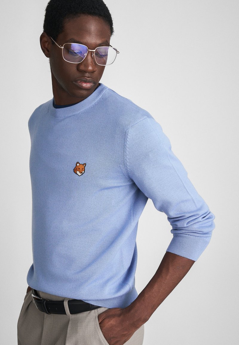 Pulover tricotat de culoare albastru deschis, cu guler tip crew, având o broderie cu un cap de vulpe pe piept. Textură moale, croială ajustată și manșete ribbate.