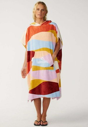 Mujer rubia que lleva un poncho de toalla con capucha multicolor con bolsillo para las manos y logo "Rip Curl", de pie descalza con chanclas negras.
