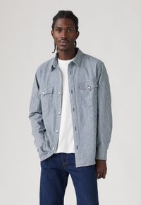 Ριγέ πουκάμισο με κουμπιά, κατασκευασμένο από ελαφρύ ύφασμα, με δύο τσέπες στο στήθος, γιακά με μυτερή άκρη και άνετη εφαρμογή. Συνδυασμένο με λευκό Τ-shirt και τζιν.