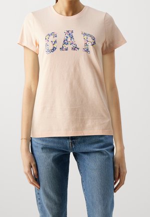 Femme portant un t-shirt rose clair à manches courtes avec le logo floral "GAP" et un jean en denim bleu sur un fond uni.
