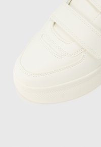 Κοντινό πλάνο στο μπροστινό μέρος ενός λευκού αθλητικού παπουτσιού με υφή στην σόλα και κλείσιμο με ιμάντα Velcro σε απλό φόντο.